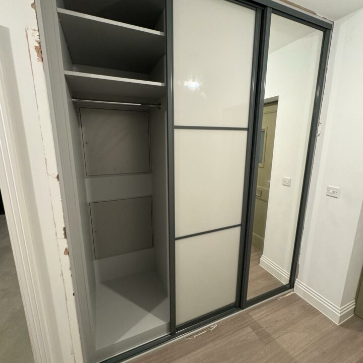 An open modern wardrobe with sliding mirrored doors, showcasing empty shelves and a hanging section. The wardrobe is in a room with wooden flooring and white walls. This fitted wardrobe integrates seamlessly into the space, providing an efficient storage solution.