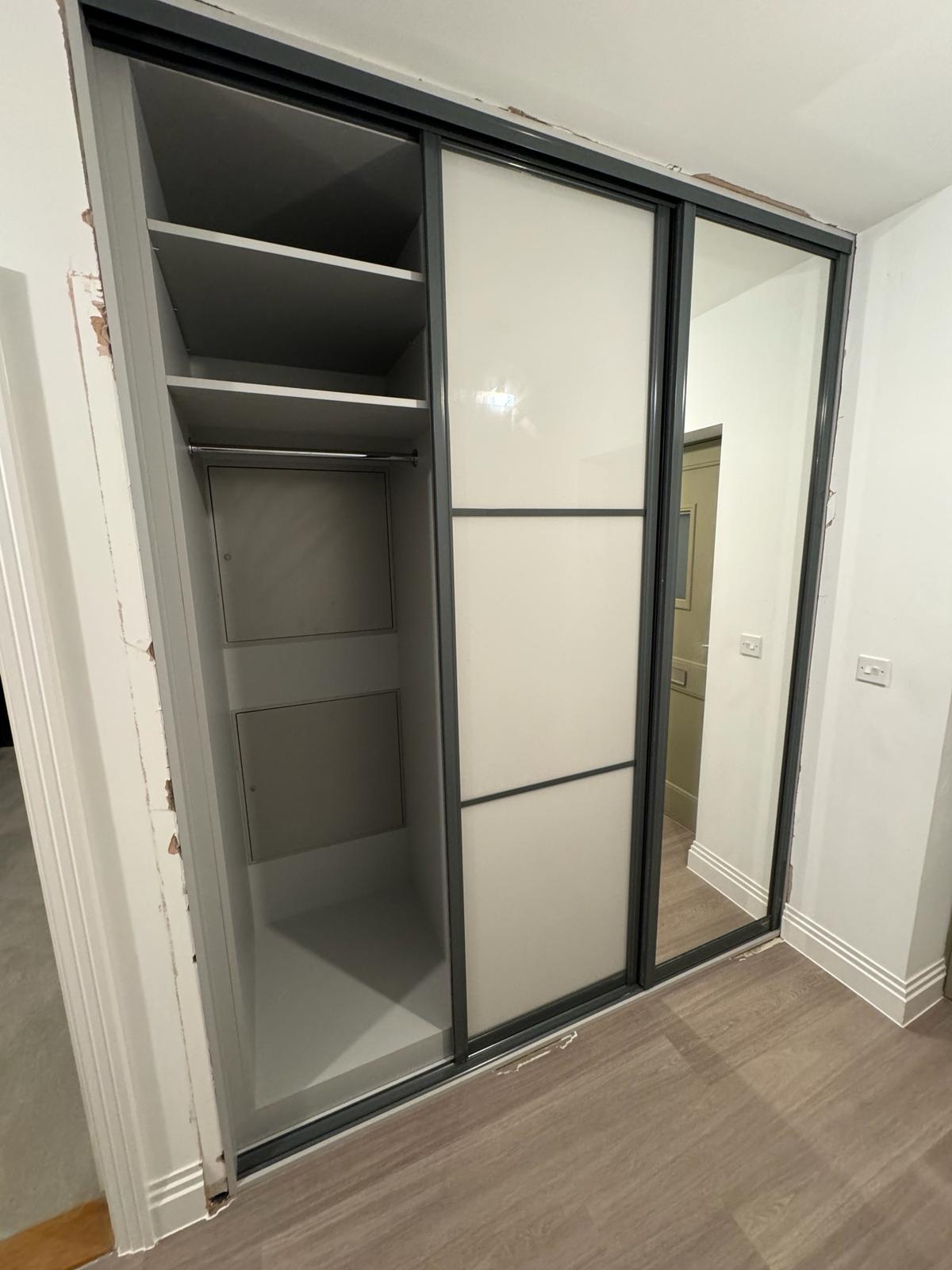 An empty built-in wardrobe with a large mirrored sliding door, partially open to reveal interior shelves and compartments, set in a room with laminate flooring and white walls. This is an essential component of our bespoke furniture collection for fitted bedrooms.