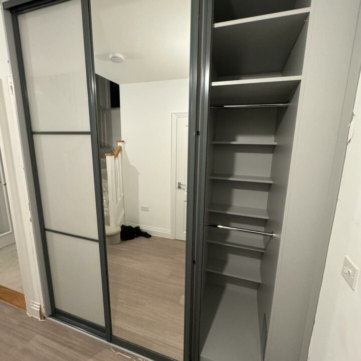 A modern fitted bedroom wardrobe with frosted glass and black frames, showing an open section with empty gray shelves and a mirrored reflection of a room.