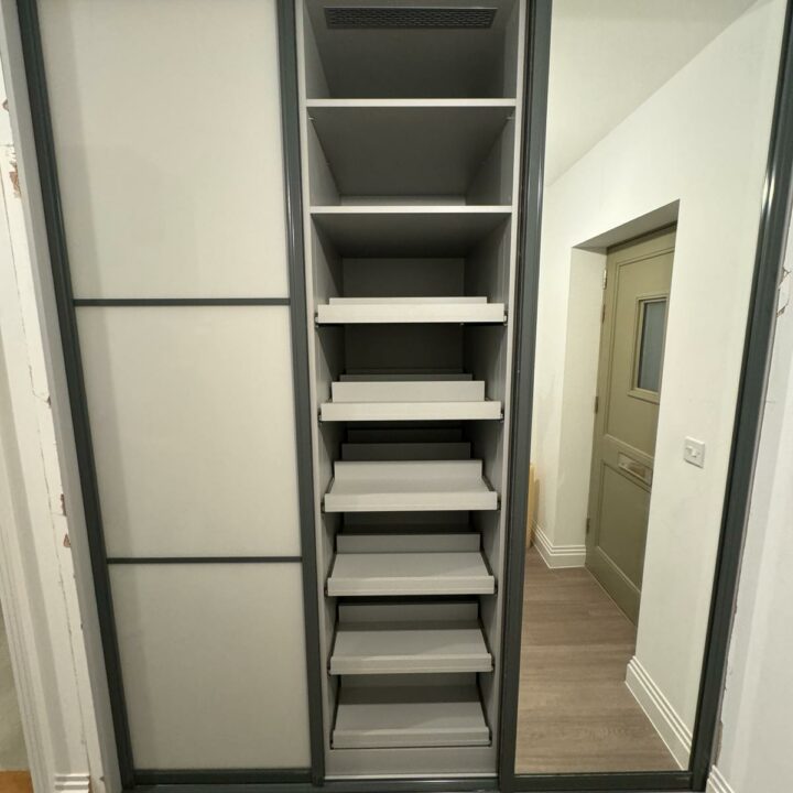 A modern, fitted wardrobe with sliding doors, featuring several white shelves organized neatly within one section, and a frosted glass segment in the other.