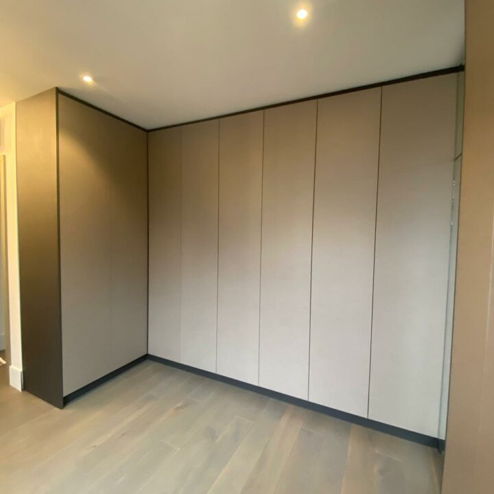A modern room with light gray walls and flooring, featuring a large, built-in wardrobe with sliding door panels. The room is well-lit with ceiling lights.