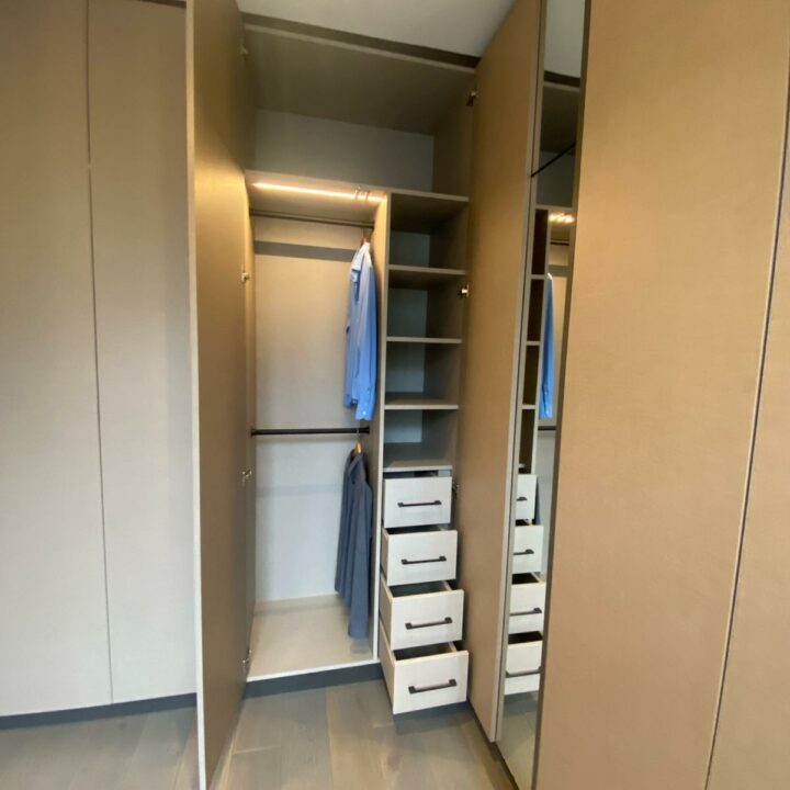 Modern walk-in closet featuring open beige fitted wardrobes, a hanging blue shirt, and several white pull-out storage drawers.