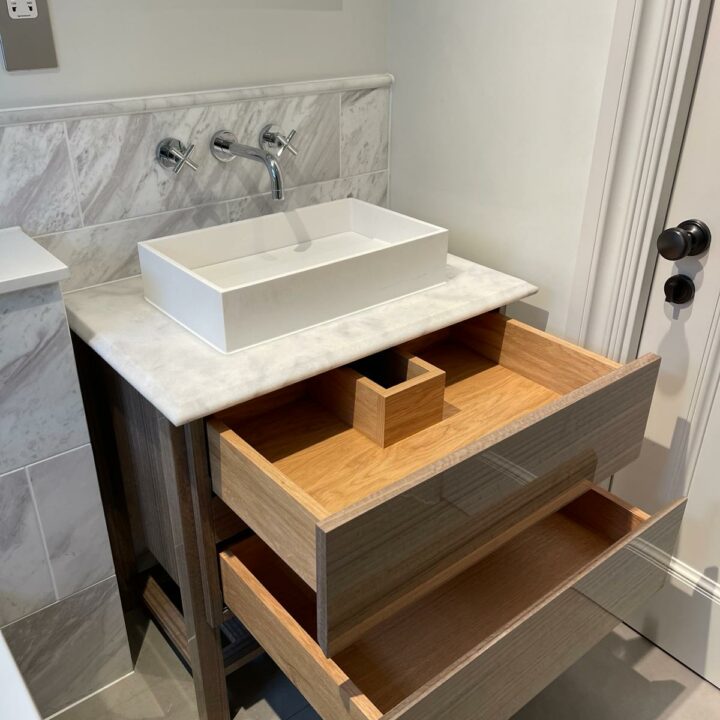 A modern bathroom vanity with a square white basin on a wooden countertop, featuring open drawers displaying organized compartments, set against a marble wall with silver fixtures and adjacent fitted wardrobes.