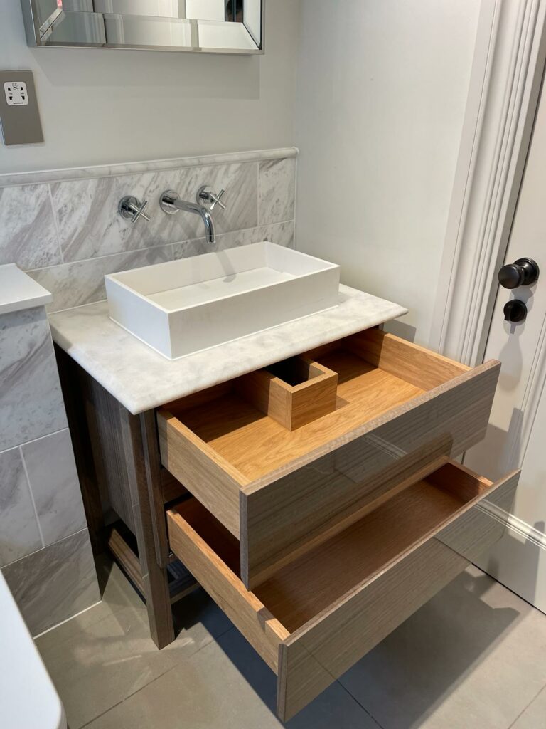 A modern bathroom vanity with a square white basin on a wooden countertop, featuring open drawers displaying organized compartments, set against a marble wall with silver fixtures and adjacent fitted wardrobes.