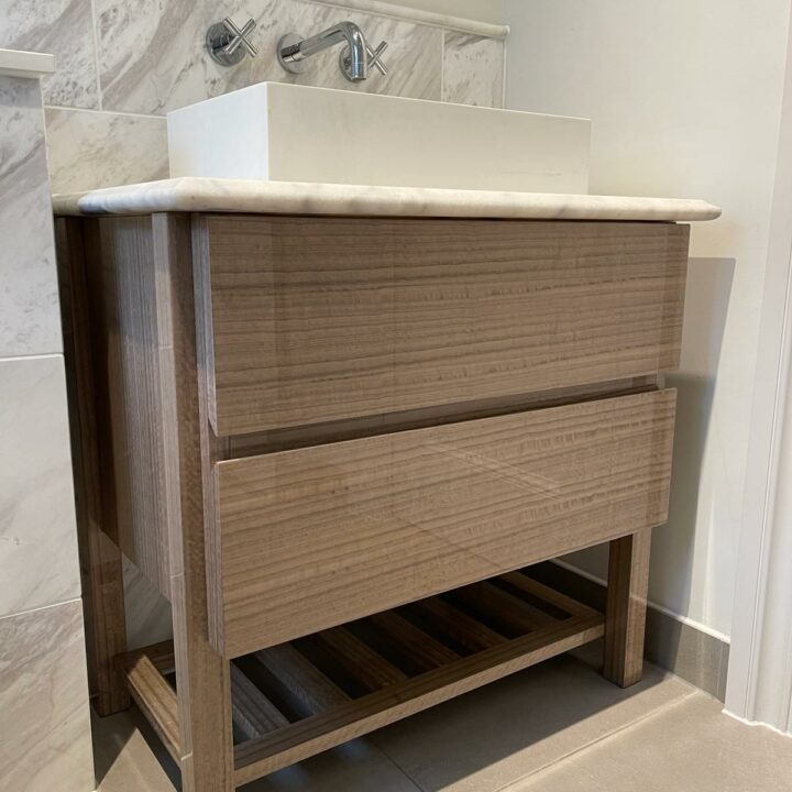 A modern bathroom vanity featuring a wooden cabinet with three drawers and an open shelf below, topped by a white sink, against a marble wall with fitted wardrobes.