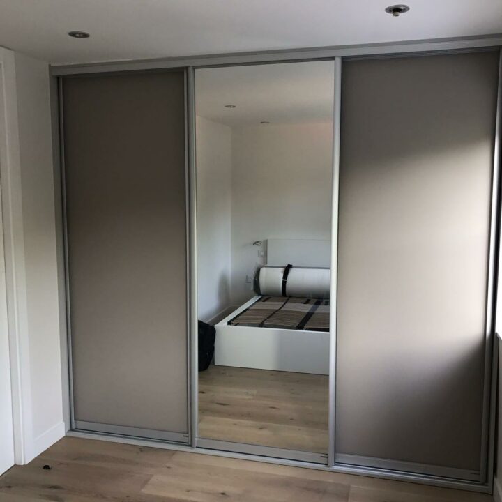 A modern bedroom reflected in large mirrored fitted wardrobes, featuring a minimalist bed and a window with natural light.