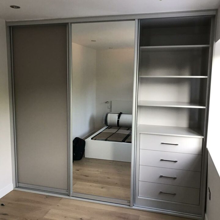 Modern bedroom featuring bespoke furniture with a sliding mirrored closet revealing built-in shelves and drawers, set against a wooden floor and neutral walls.