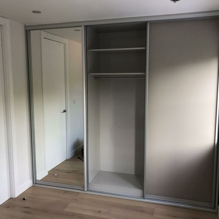A modern, empty fitted wardrobe with mirrored sliding doors, featuring several shelves and a hanging rod, in a room with hardwood floors and natural light.