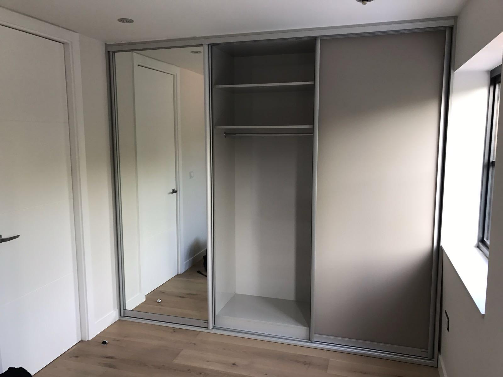 A modern, empty fitted wardrobe with mirrored sliding doors, featuring several shelves and a hanging rod, in a room with hardwood floors and natural light.