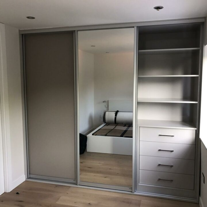 A modern bedroom featuring a large fitted wardrobe with sliding doors, a mix of mirrored and opaque panels, and an interior organizer with shelves and drawers. The flooring is wooden.