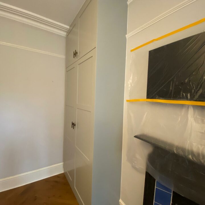 A tall white His and Hers fitted wardrobe with brass handles in the corner of a room with wooden flooring, white walls, crown molding, and a partially visible black fireplace with blue tiles, covered with protective
