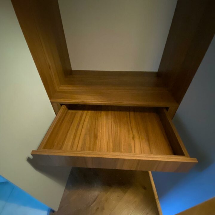 An open wooden shelf mounted on a wall in a corner, with a blue glow from ambient lighting at the bottom, positioned over a wooden floor, adjacent to His and Hers fitted wardrobes.