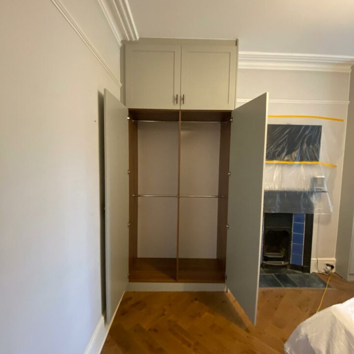 A His and Hers fitted wardrobe in an empty room with open doors, featuring an upper storage compartment, a bar for hanging clothes, and a backdrop of a covered fireplace.