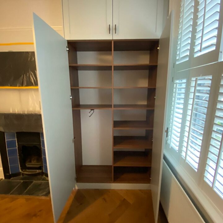An open, empty fitted wardrobe with several shelves, designated as "His and Hers", situated in a well-lit room with white shutters on the windows and a closed fireplace covered with black plastic.