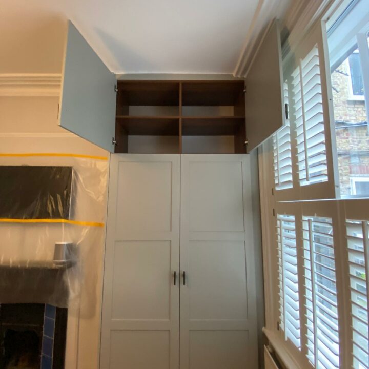 A well-organized room featuring a large white "His and Hers Fitted Wardrobes" with upper wooden shelves against a white wall. A window with blinds is on the left, and protective plastic
