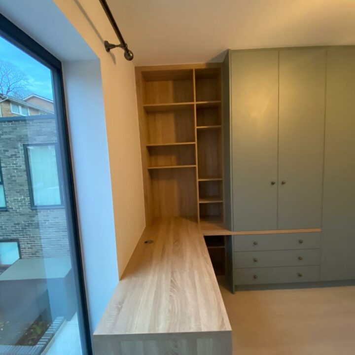 Interior view of a modern room featuring a long wooden bench, built-in shelving unit to the left, and wardrobes with built-in desks to the right, with a large window showing an exterior