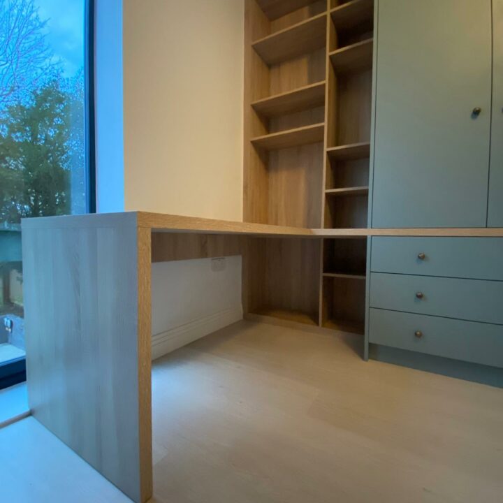 An empty room corner with a built-in wooden desk, bookcase, and wardrobes, all finished in light wood tones. A large window provides natural light, contrasting with the pale walls and floor