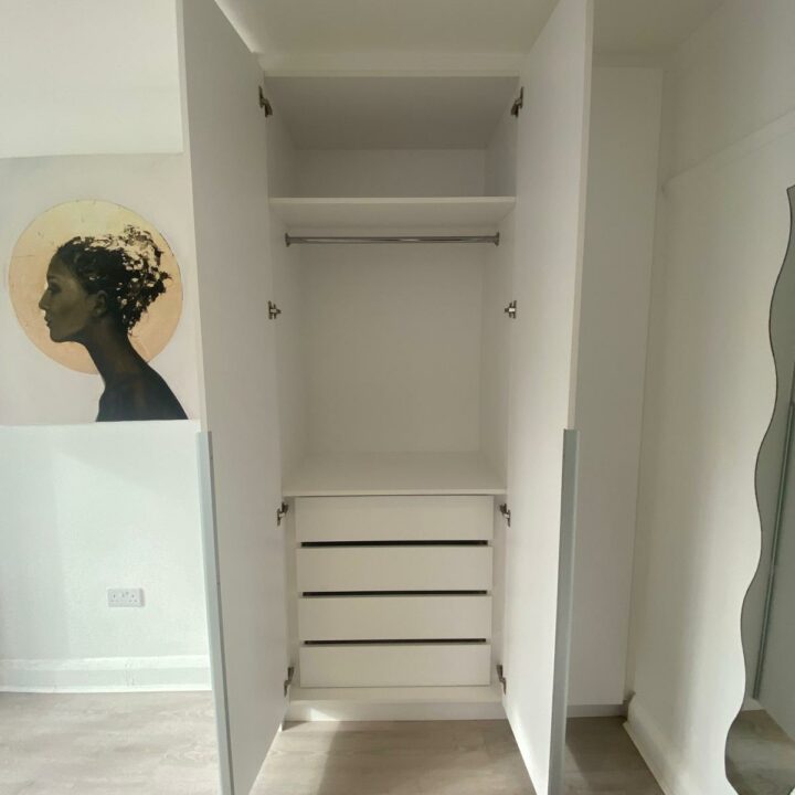 An open, built-in wardrobe with hinged doors in a room with light-colored walls and a wooden floor. The wardrobe features an empty shelf and several drawers, and there are artistic paintings on the adjacent