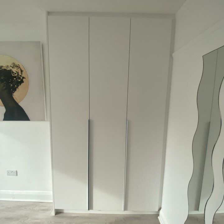 A crisp white fitted closet with three vertical hinged doors situated in a room with light gray flooring, and partial views of an ornate mirror and a dark-toned painting.