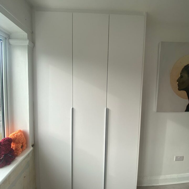 A white three-door wardrobe with hinged doors in a room corner beside a window, with a coral decoration on the windowsill and a round art piece on the adjacent wall.