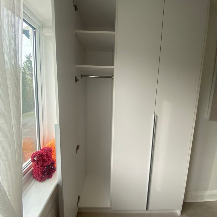 A white, narrow wardrobe with hinged doors open, revealing shelves inside, placed beside a sunny window with translucent curtains in a room with wooden flooring. A pink basket sits by the window.