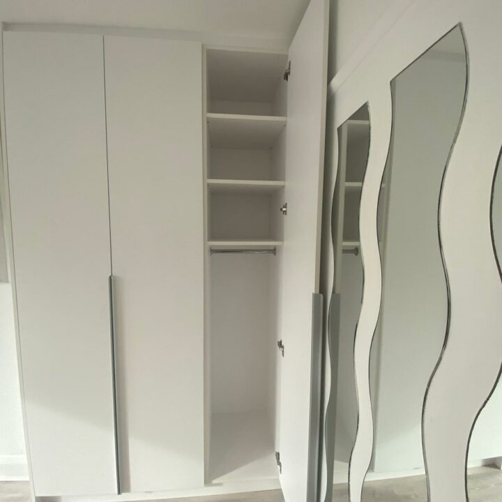 Interior of a room showing an open bespoke wardrobe with empty shelves and a curvy mirror on the right side.