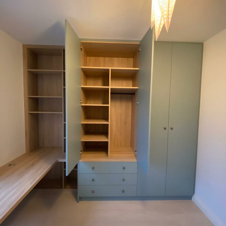 A well-lit, empty walk-in closet featuring a combination of open shelving, drawers, wardrobes with built-in desks, and a hanging area, all crafted in light wood tones against muted