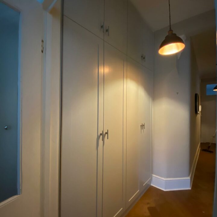 A dimly lit hallway featuring a large, built-in His and Hers Fitted Wardrobes with sleek handles, a wooden floor, and a hanging pendant light emitting a warm glow.
