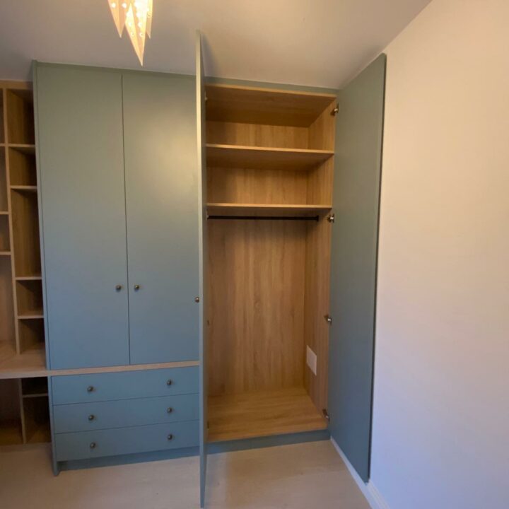 An open wardrobe with built-in desks and wooden shelves, painted in light blue, situated in a room with beige walls and a wooden floor.