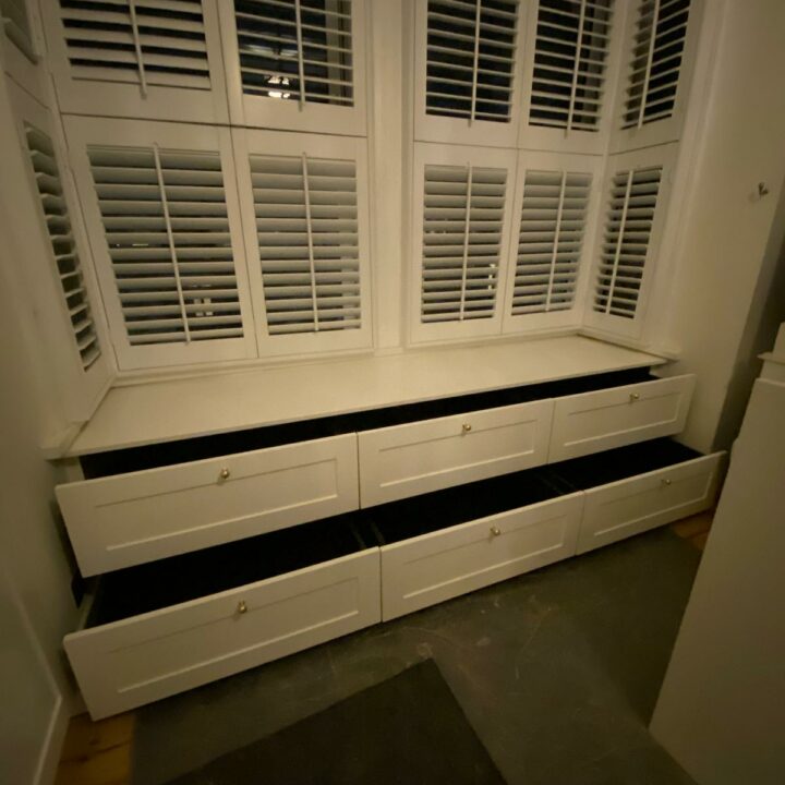 A dimly lit image showing a bespoke seating area with storage drawers under large windows covered by white shutters.