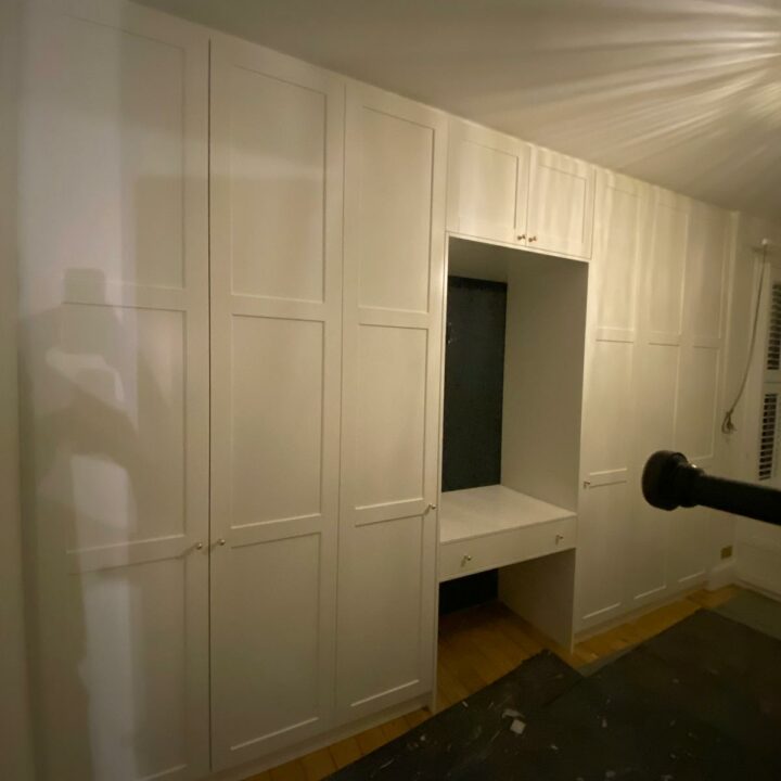 A white fitted wardrobe with multiple hinged doors and a central open nook with a bench, set in a room with dark hardwood floors and a white, draped ceiling.