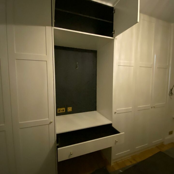 A white bespoke built-in wardrobe with an open cabinet and a drawer in a dimly lit room, showing an empty shelf and a black back panel.