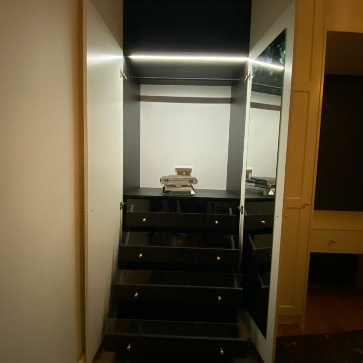 A dimly lit hallway with black stairs leading up to a small, illuminated landing where a stack of books sits under a bright light, flanked by fitted wardrobes.