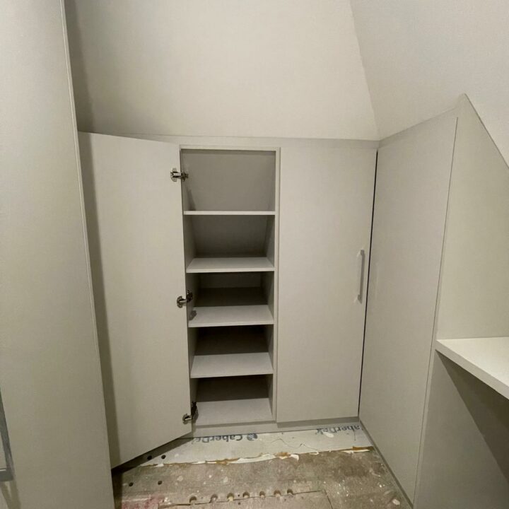 A small, under-construction closet space with white bespoke furniture and shelves under a slanted ceiling, featuring an open cabinet door and visible construction debris on the floor.