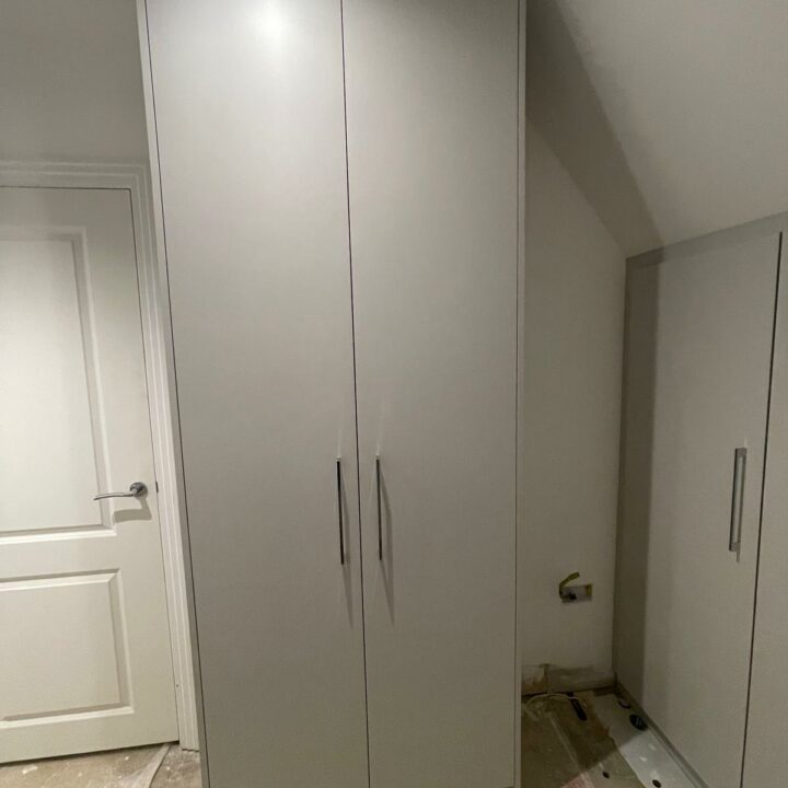 A large, grey fitted wardrobe with two doors, installed in a corner of a room with white walls and a wooden floor next to another door.