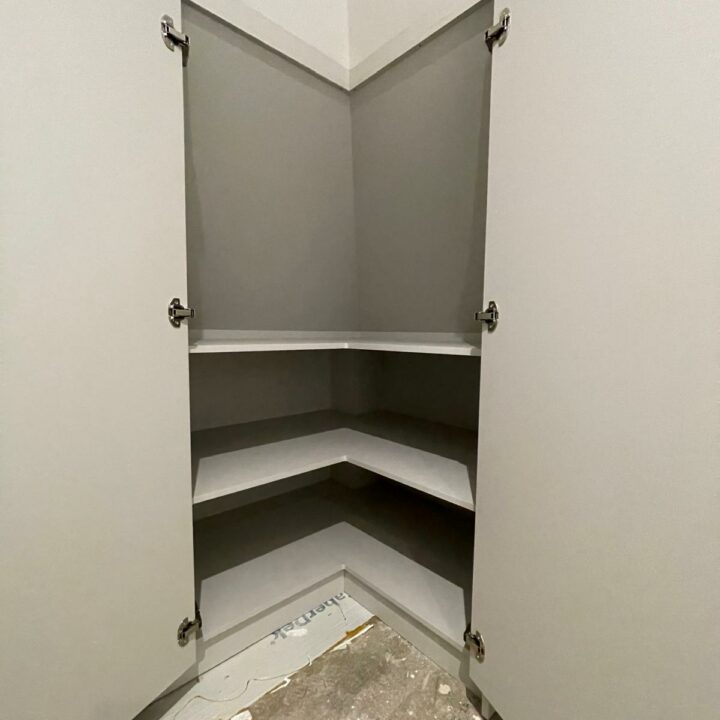 A newly constructed empty walk-in wardrobe corner with two white shelves, gray interior walls, and remnants of construction material on the floor.
