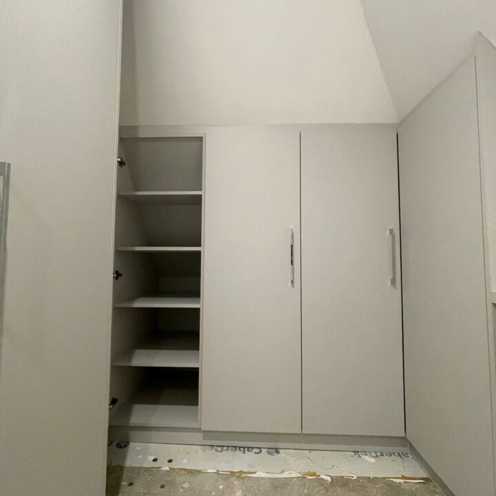A narrow closet with open shelving on the left and hinged door wardrobes on the right, located in a room with unfinished flooring and white walls.