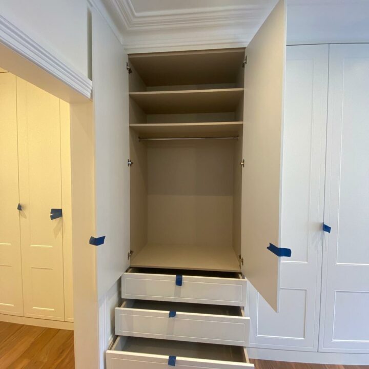 An open fitted wardrobe with multiple empty shelves above three drawers, located in a room with wooden flooring and white paneled walls, each drawer and shelf door marked with blue tape, ready to paint.