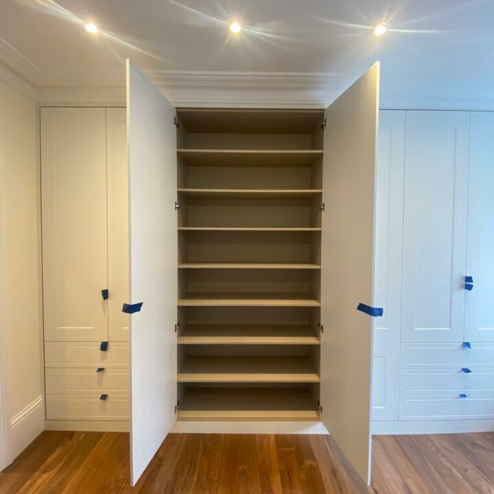 An open fitted wardrobe with multiple shelves and white doors, showing a sleek modern design with hardwood flooring and recessed ceiling lights.