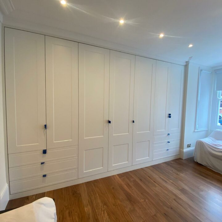 A spacious room featuring a large built-in white wardrobe with multiple hinged door wardrobes and dark handles, a wooden floor, and recessed ceiling lights. A bed is partially visible on the right.