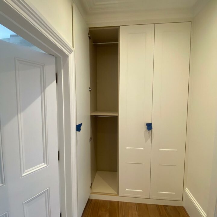 A corner view of a newly installed white fitted wardrobe with multiple doors, adjacent to an open white door leading to a wooden-floored room. Blue protective film covers the wardrobe's handles.