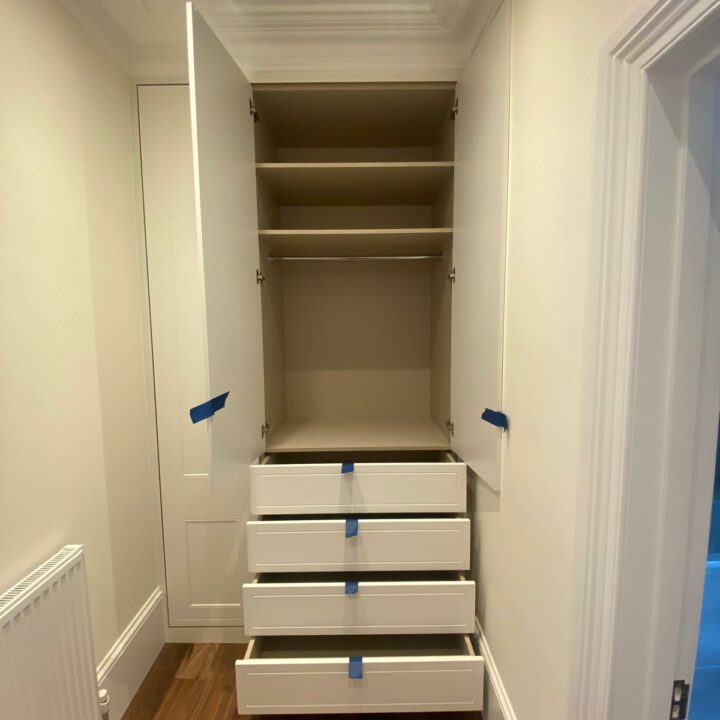A narrow, newly constructed walk-in wardrobe with multiple white drawers with blue tape handles, leading up to a tall, open-shelved closet, in a hallway with beige walls and white doors.