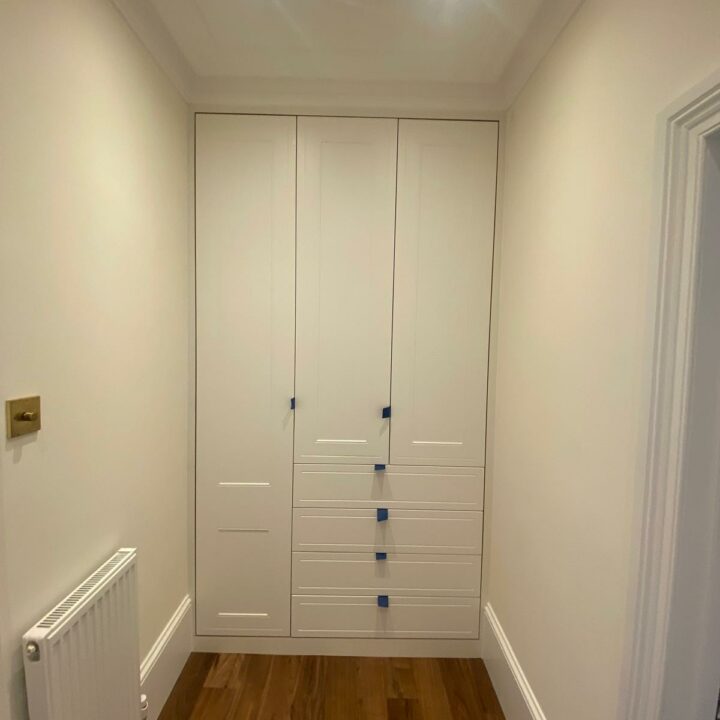 A modern hallway featuring sleek wooden flooring and a white paneled double door with blue protective tape on the handles. The walls are painted white, and there are three ceiling spotlights. Along one side, fitted wardrobes blend seamlessly with the decor.
