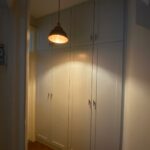 A warmly lit hallway featuring a large, white His and Hers fitted wardrobe with closed doors, and a hanging bronze pendant light illuminating the space.