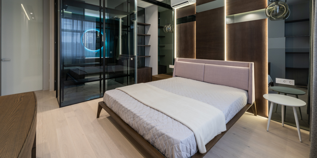 Modern bedroom with a double bed, light-colored bedding, and wooden furnishings. The room features mirrored sliding doors, a small round bedside table, bespoke fitted furniture with built-in shelving, and contemporary lighting fixtures. The decor is minimalist with a focus on clean lines.