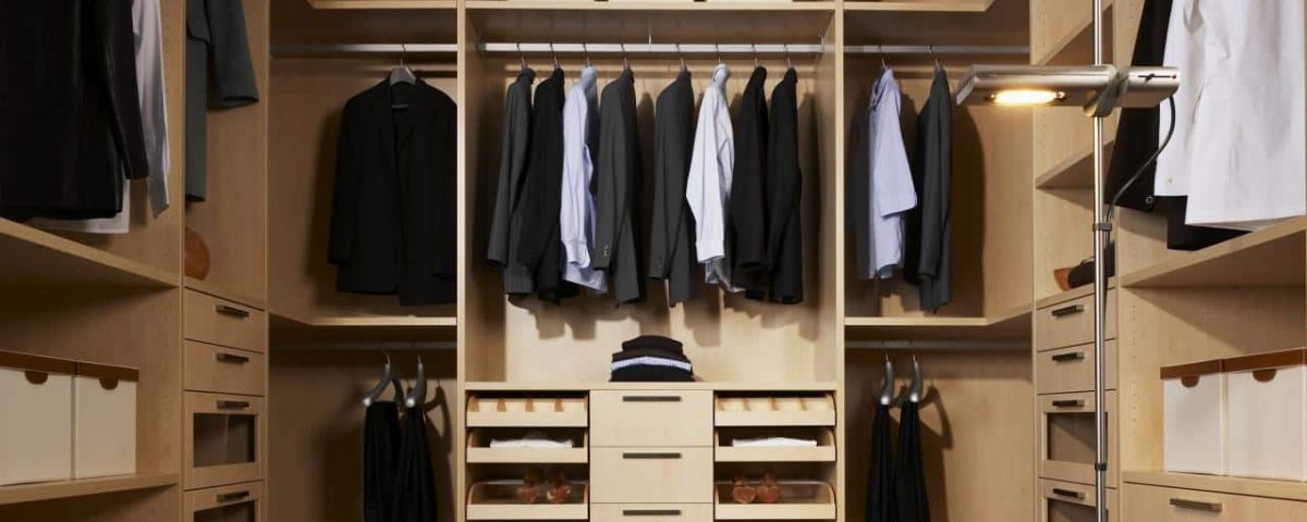 A neatly organized walk-in closet with bespoke fitted furniture, including wooden shelves and a variety of clothing items hanging. Suits and jackets are on hangers, while shirts and pants are folded on shelves. There's also a central drawer unit and a standing lamp providing light.