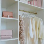 A neatly organized closet with white shelves showcases smart storage solutions, holding several items, including pink handbags, boxes, and pink gift bags. On a gold hanging rod, there are three clothing items: a beige ruffled sweater, a white blouse, and a yellow sweater. A small basket is on the bottom shelf.