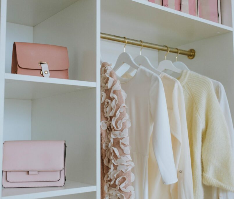 A neatly organized closet with white shelves showcases smart storage solutions, holding several items, including pink handbags, boxes, and pink gift bags. On a gold hanging rod, there are three clothing items: a beige ruffled sweater, a white blouse, and a yellow sweater. A small basket is on the bottom shelf.