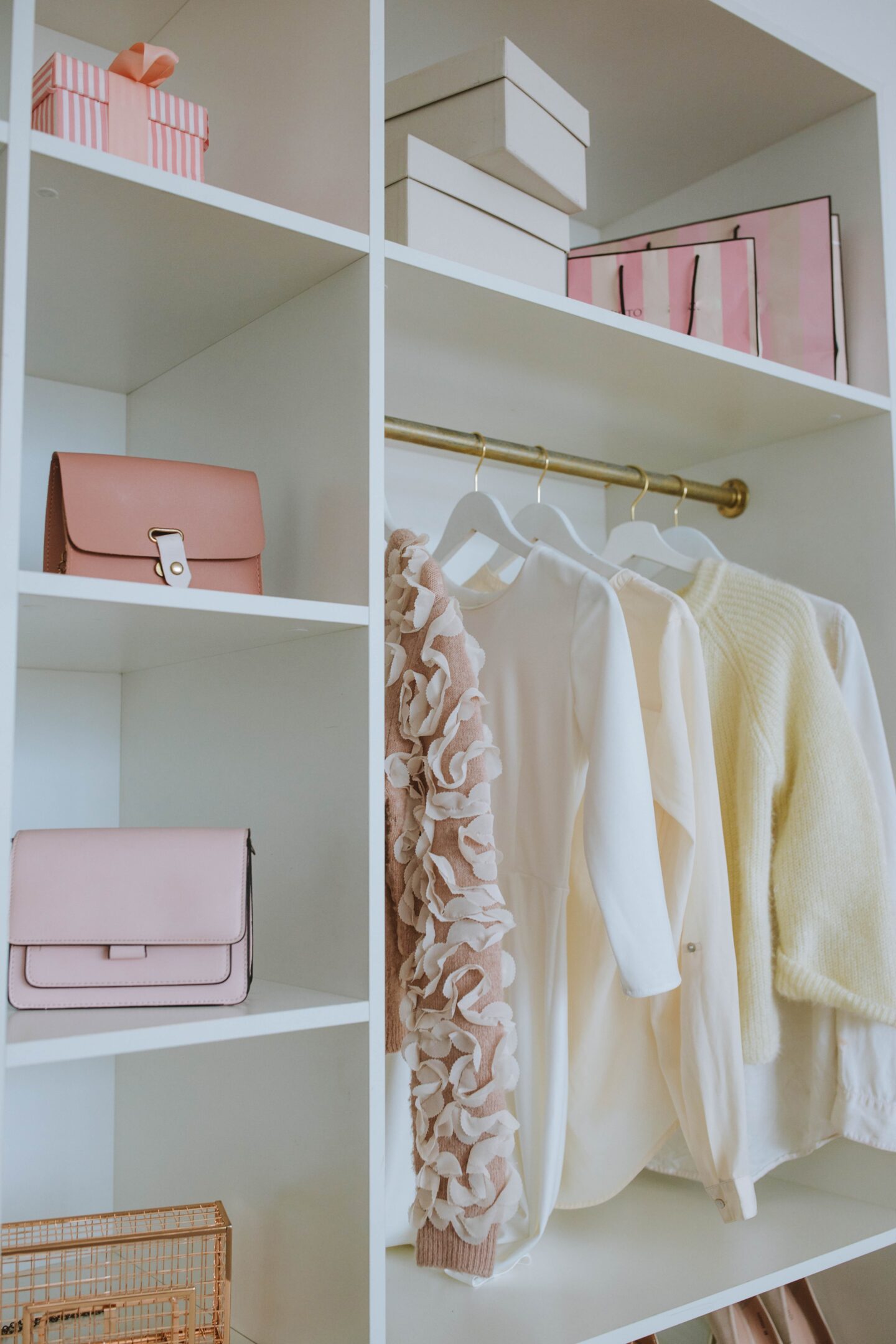 A neatly organized closet with white shelves showcases smart storage solutions, holding several items, including pink handbags, boxes, and pink gift bags. On a gold hanging rod, there are three clothing items: a beige ruffled sweater, a white blouse, and a yellow sweater. A small basket is on the bottom shelf.