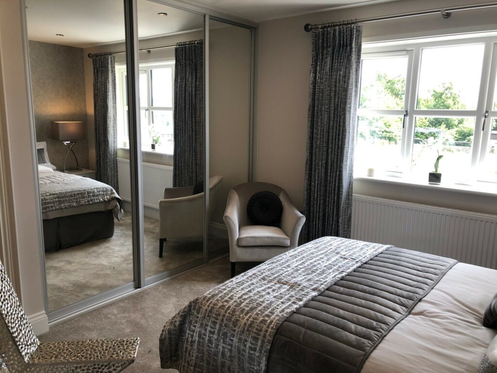 A modern bedroom features a large bed with gray linens, a white armchair, and mirrored closet doors reminiscent of IKEA Pax alternatives. Two windows with gray curtains let in natural light, illuminating the carpeted floor and a small side table with a lamp.